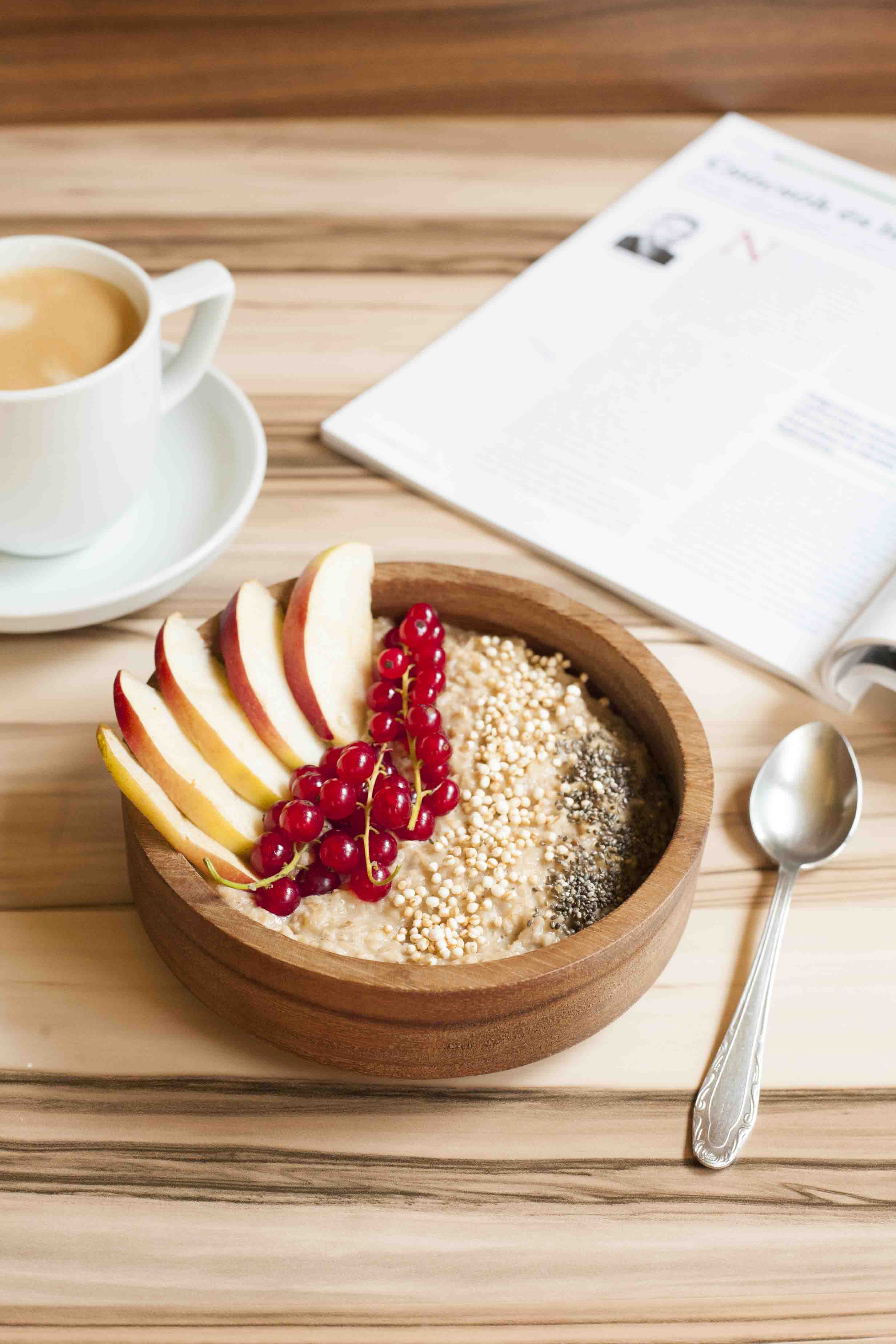 A wooden bowl of oatmeal topped with apple slices, red currant, chia seeds and puffed quinoa