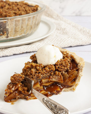 A slice of apple pie on a white plate with brown sauce, apple slices and crumbly topping. There is a fork taking the first bite.