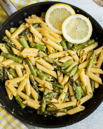 Frying pan from above with penne pasta, asparagus pieces and 2 lemon slices.