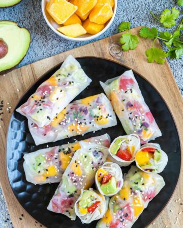 Fresh rice paper wraps filled with chopped tomato, mango, and avocado placed on a black plate. 4 half rolls and 6 whole rolls. Leftover ingredients are next to it.