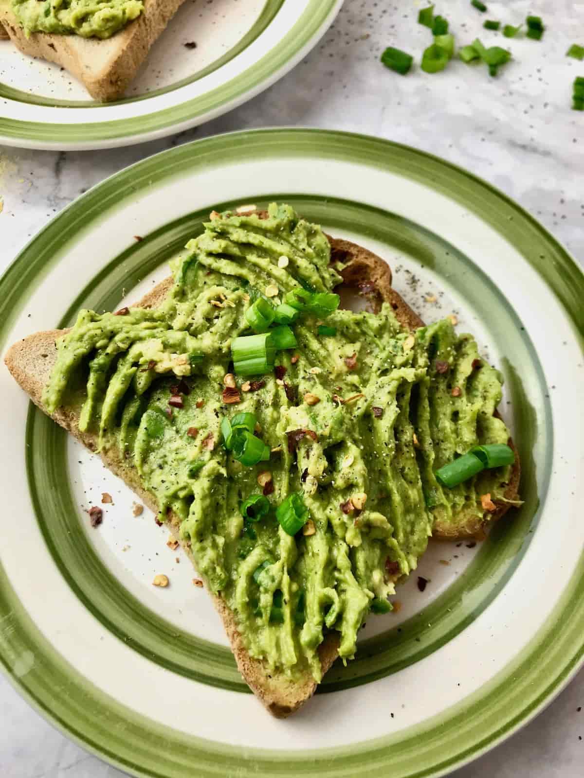 A slice of toast on a white green plate topped with mashed avocado, scallion and chili flakes.