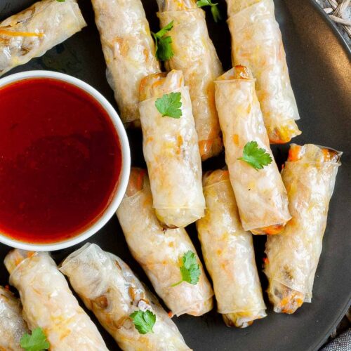 Black plate with crispy looking light brown egg rolls. A small white bowl is with a red dipping sauce. The rolls are sprinkled with parsley leaves.