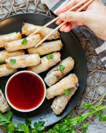 Black plate with crispy looking light brown egg rolls. A small white bowl is with a red dipping sauce. The rolls are sprinkled with parsley leaves.