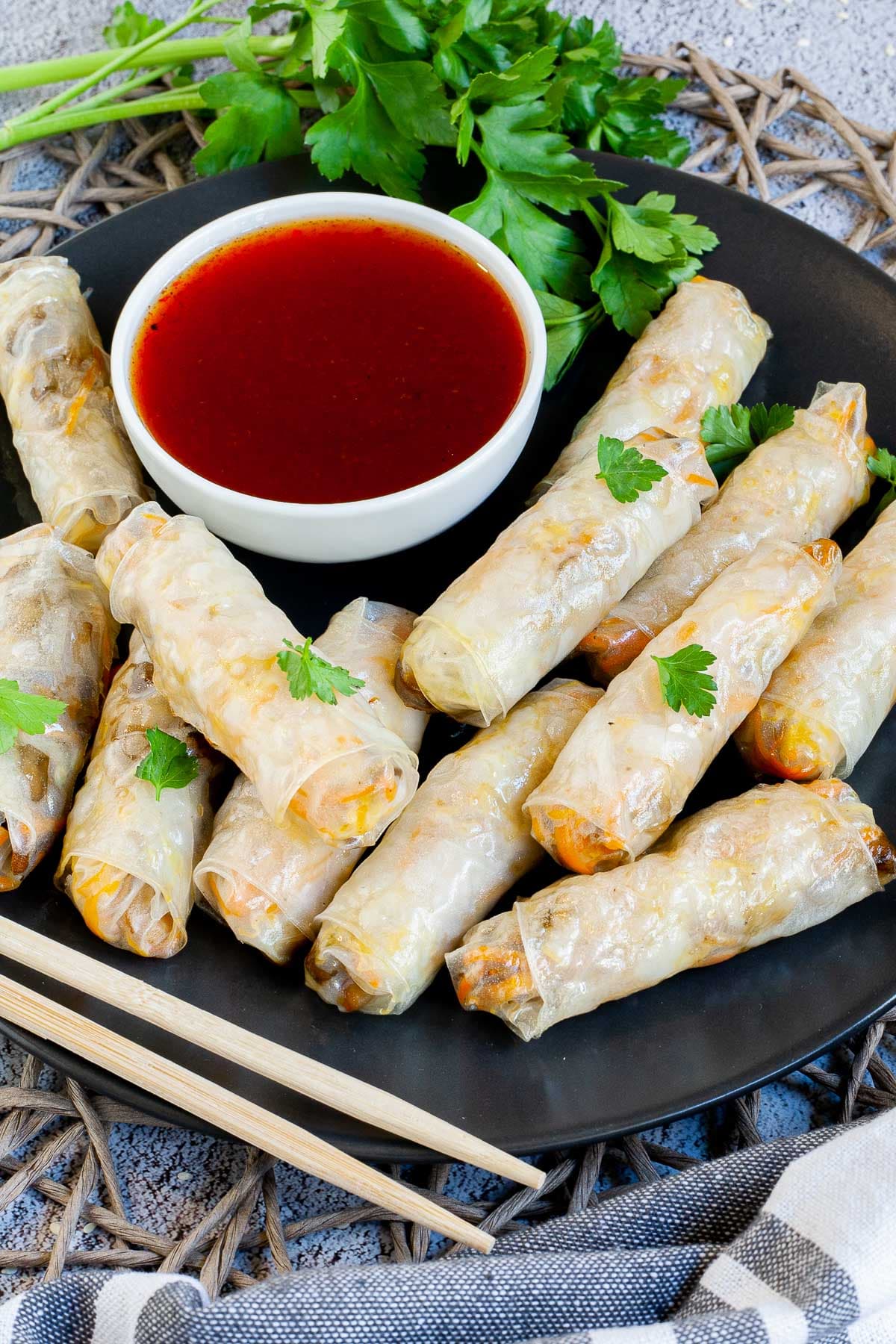 Black plate with crispy looking light brown egg rolls. A small white bowl is with a red dipping sauce. The rolls are sprinkled with parsley leaves.