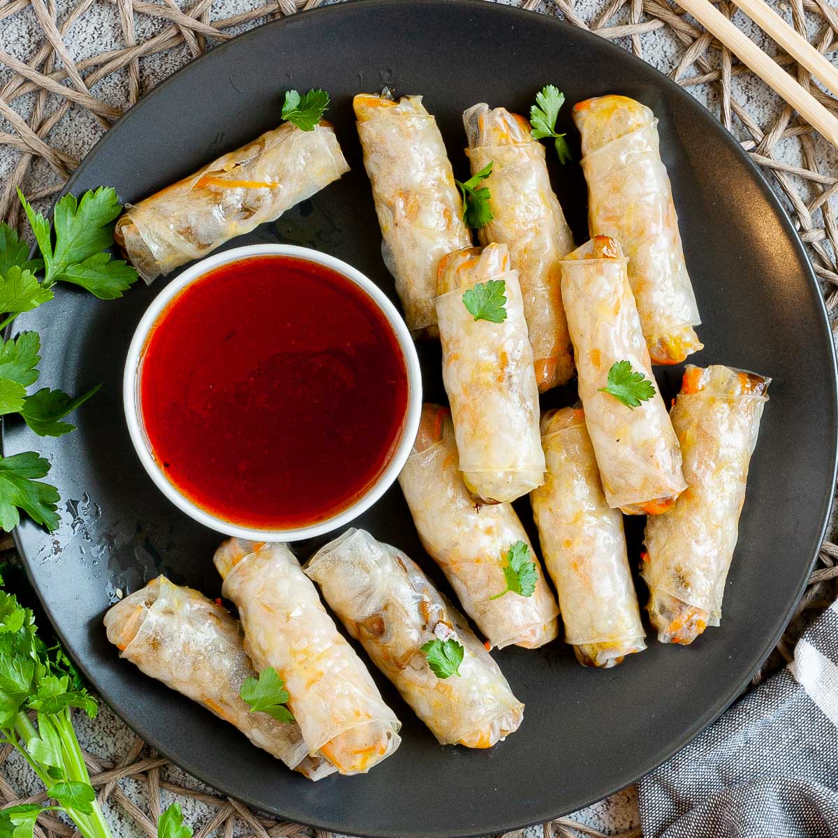Black plate with crispy looking light brown egg rolls. A small white bowl is with a red dipping sauce. The rolls are sprinkled with parsley leaves.