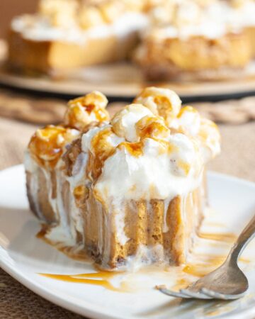 One slice of amber brown cheesecake with whipped cream, caramel sauce and chopped nuts on top is served on a white plate. One bite is missing from the front. The whole cake is in the background.