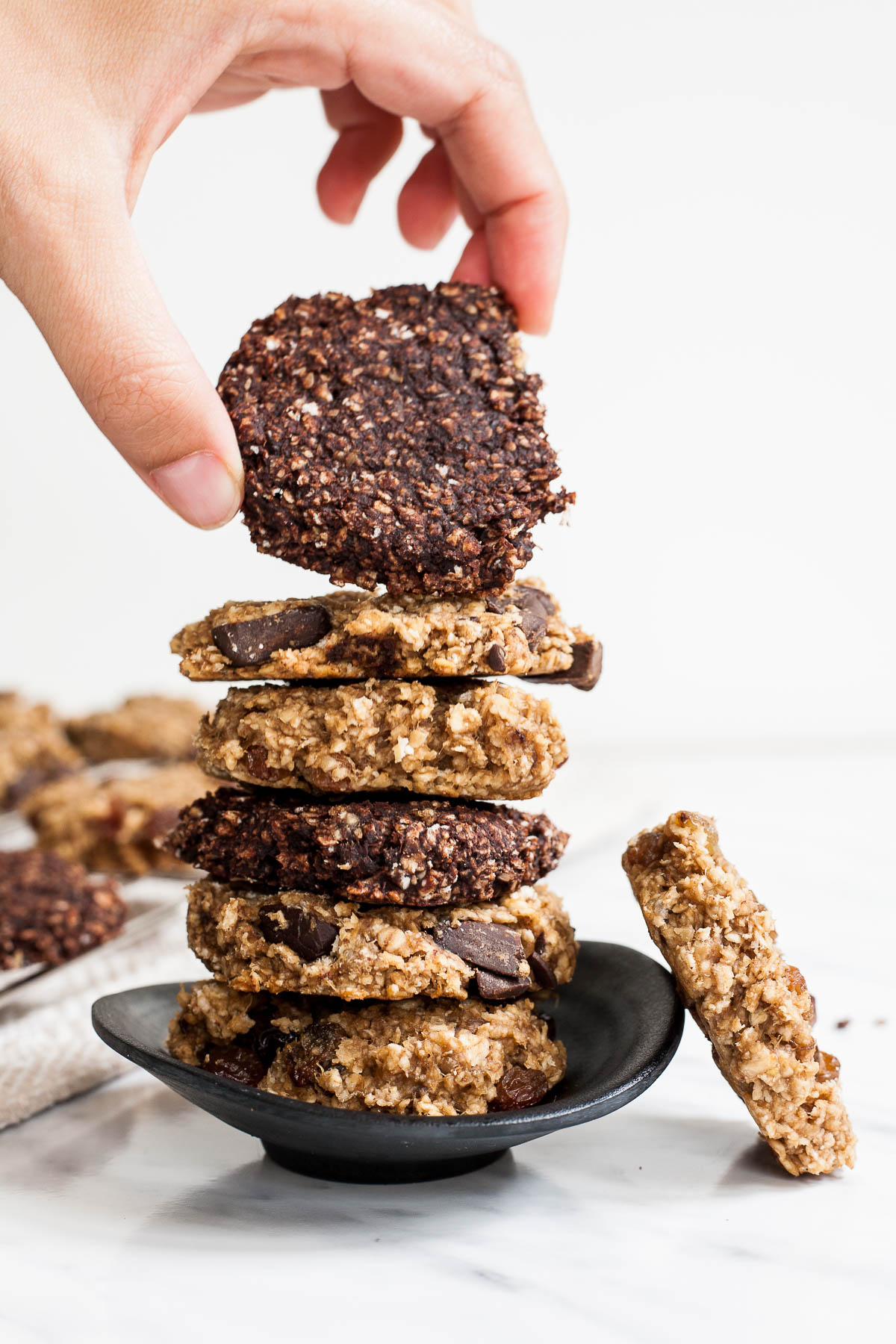 Stack of brown and dark brown oatmeal chocolate cookies on a small black bowl. A hand is taking the top cookie. 