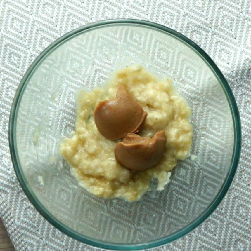 A glass bowl with mashed bananas and peanut butter scoops.