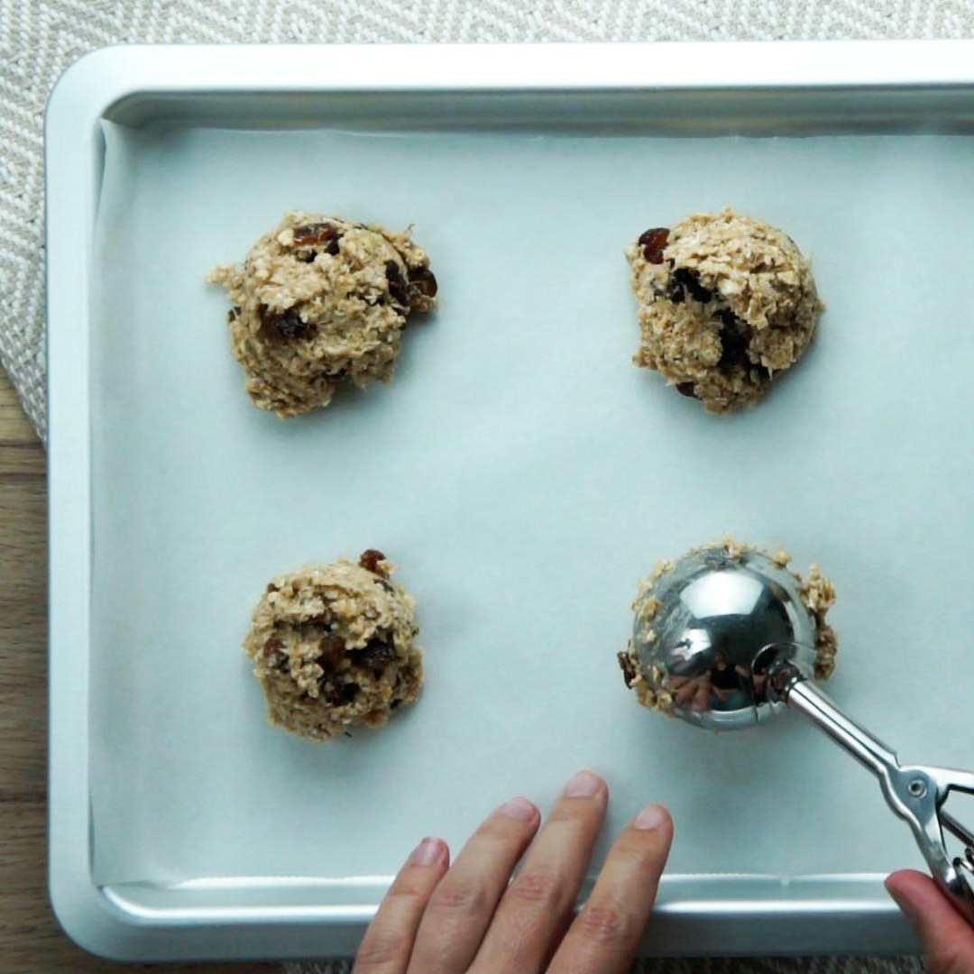 A hand is using an ice cream scoop to portion oatmeal cookies to a silver baking sheet.