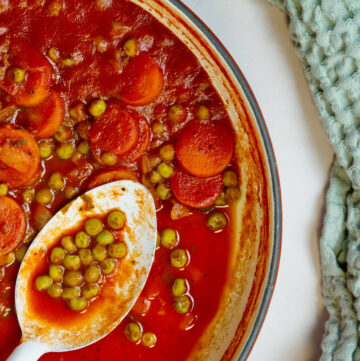 Vibrant red stew with peas and carrots in a white red enameled Dutch oven. A spoon is inside taking some peas.