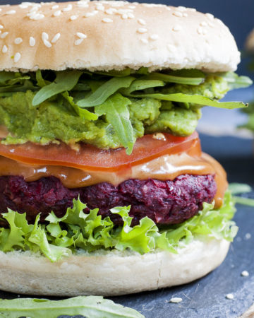 Close-up of a beet burger with layers like burger bun, lettuce, purple patty, tomato slice, green puree, arugula and bun on the top
