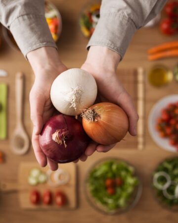A hand is holding three different type of onions. More ingredients are in the background.