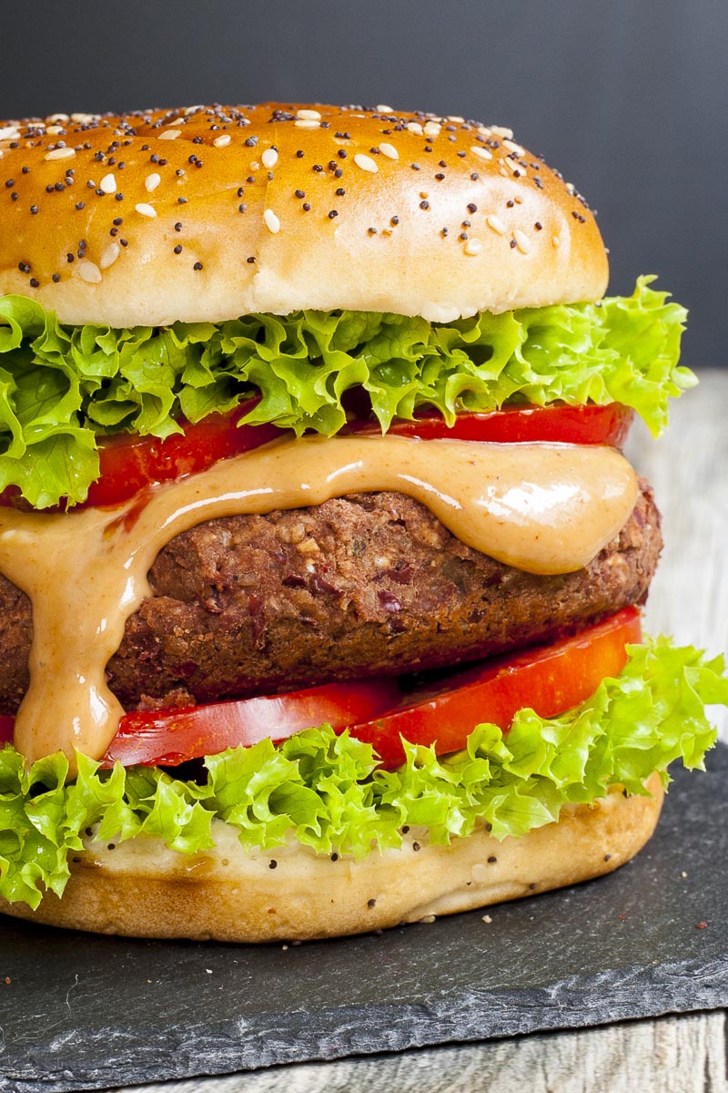 A burger on a black board with lettuce, tomato, orange sauce and a dark brown burger patty.