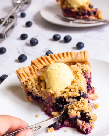 One slice of blueberry pie on a white plate with vibrant purple berries, crumble topping and a scoop of yellow ice cream. Another plate with a slice is in the background. Fresh blueberries are scattered around.