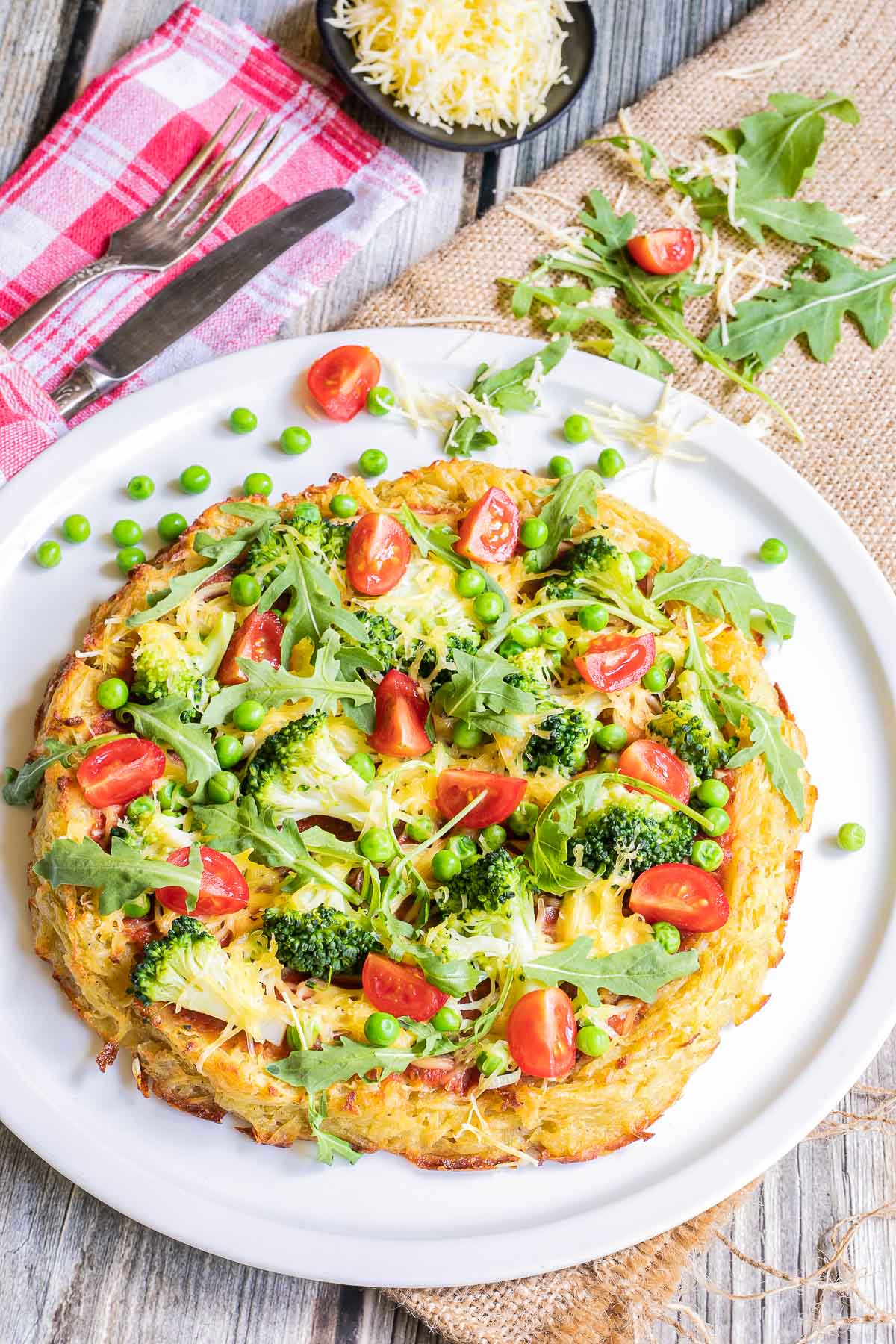 Hash brown crust pizza with topped with tomato sauce, broccoli, peas cherry tomatoes, arugula, and shredded melted cheese. Served on a white plate.