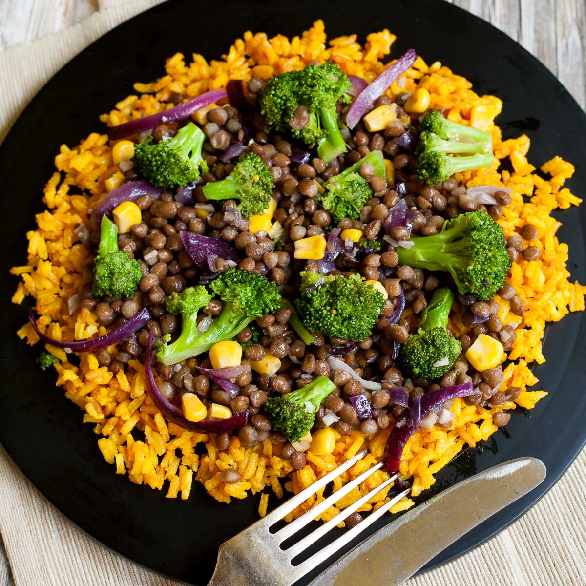 Black plate with vibrant yellow rice and a broccoli lentil stir fry with red onion slices and sweet corn.
