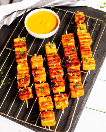 Yellow-brown tofu cubes on wooden skewers are criss cross over each other on a grilling rack placed on a black board. A small white bowl is next to them with a yellow dipping sauce.