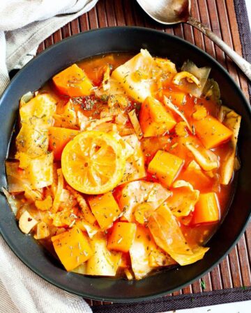 Black plate from above with red tomato-based soup and chopped vegetables like cabbage, orange sweet potato, celery stalk slices, topped with lemon slices and dried green herbs