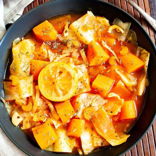 Black plate from above with red tomato-based soup and chopped vegetables like cabbage, orange sweet potato, celery stalk slices, topped with lemon slices and dried green herbs