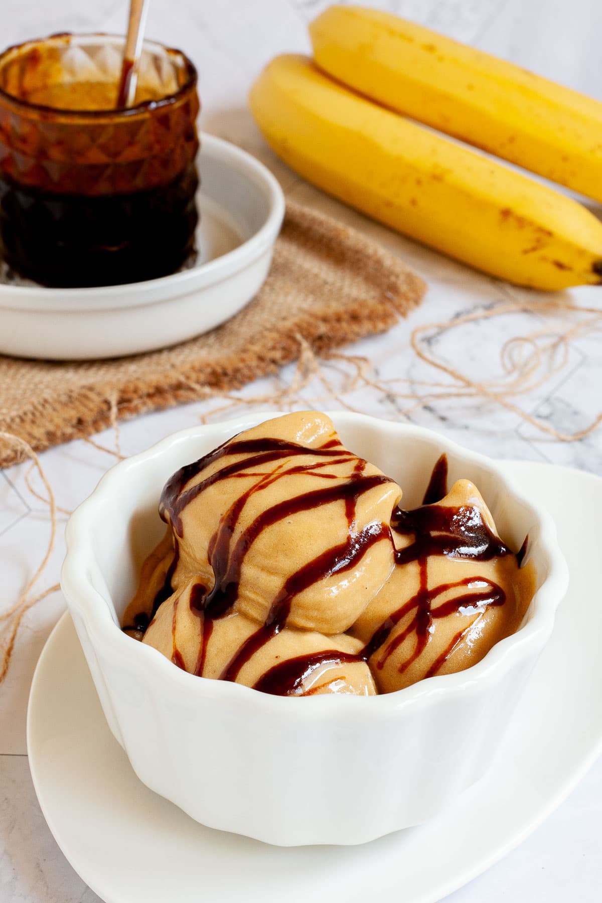 A white bowl of caramel colored ice cream with dark brown caramel drizzle on top. A glass jar of caramel sauce and 2 bananas are in the background.