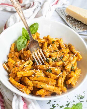 A white bowl with twisted short pasta in a creamy orange sauce topped with grated cheese and chopped green herbs. A fork is placed in the middle.