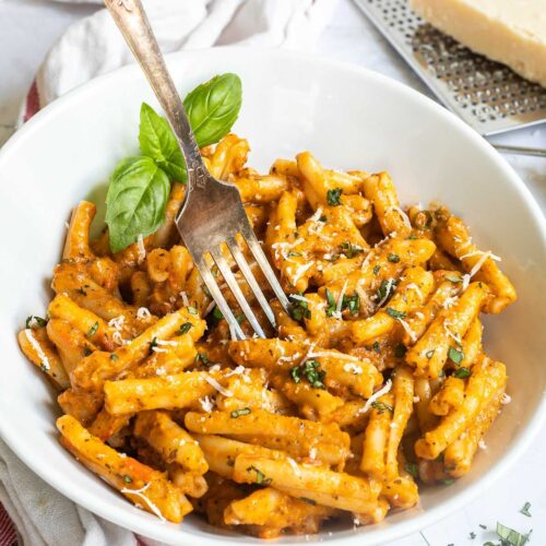 A white bowl with twisted short pasta in a creamy orange sauce topped with grated cheese and chopped green herbs. A fork is placed in the middle.