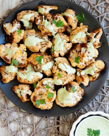 Black plate full of roasted cauliflower florets drizzled with a light green creamy sauce and sprinkled with fresh parsley. a small white bowl with the green creamy sauce is next to it.