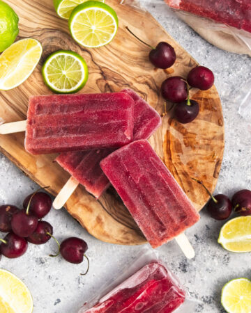 Vibrant purple popsicles on a wooden board. Cherries and lime slices are around them.