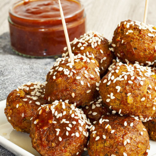 White plate with several brownish-redish balls on top of each other sprinkled with sesame seeds. In the background a small glass jar with red sauce.