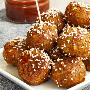 White plate with several brownish-redish balls on top of each other sprinkled with sesame seeds. In the background a small glass jar with red sauce.