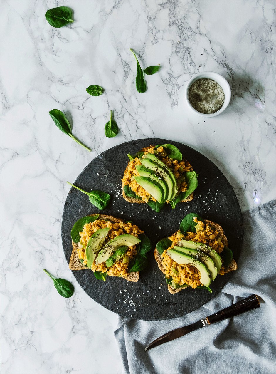 Black plate with 3 slices of bread topped with a yellow crumbly spread and avocado slices