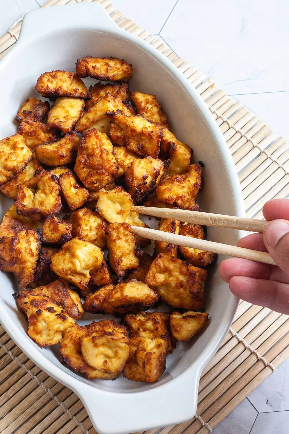 White bowl with yellow-brown tofu bites. A hand is holding chopsticks and taking one.