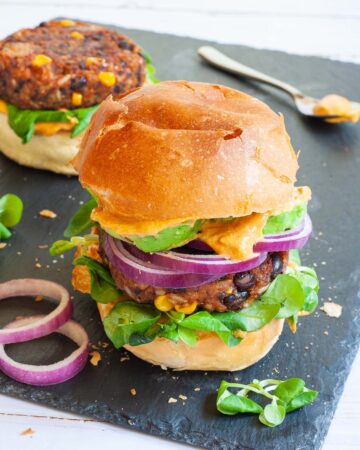 A burger bun with yellow sauce and green leaves is topped with a thick brown burger patty with visible sweet corn and black bean pieces, then red onion and avocado slices drizzled with yellow sauce again. It is covered with a top bun.