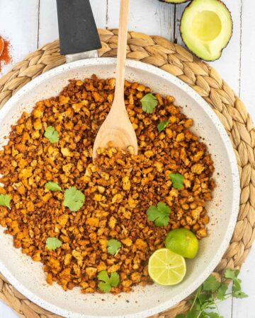 A white frying pan with brown crispy tofu crumbles sprinkled with freshly chopped cilantro. Lime slices and a wooden spatula are added. Avocado and more spices are next to the pan.