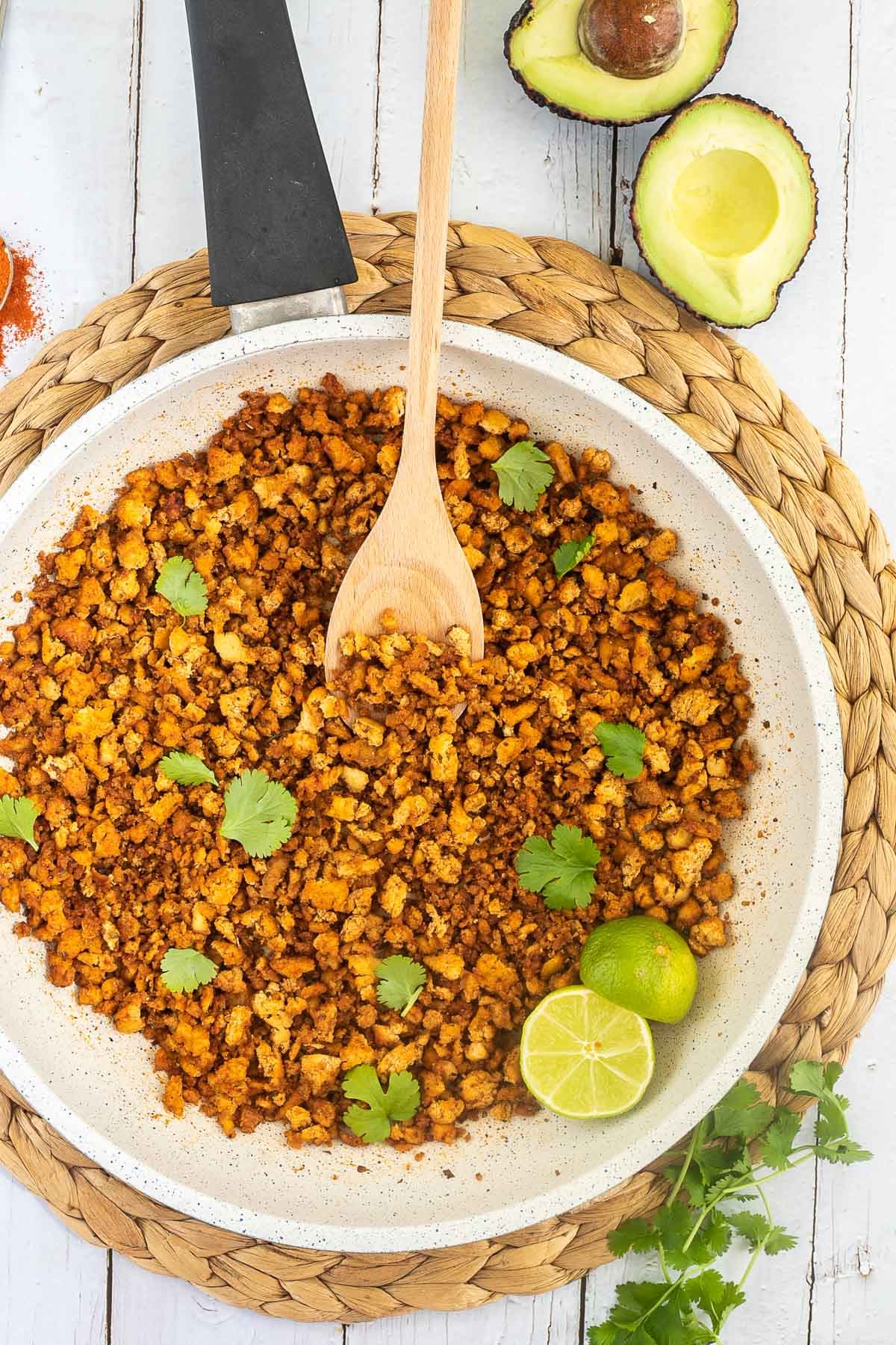 A white frying pan with brown crispy tofu crumbles sprinkled with freshly chopped cilantro. Lime slices and a wooden spatula are added. Avocado and more spices are next to the pan.