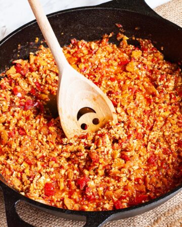 A black cast iron skillet from above with yellow brown pieces in a red chunky sauce. There is a wooden spatula with a smiley face on it placed across the sides.