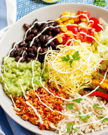 A large white plate in the middle with colorful ingredients like black beans, corn, shredded cheese, bell pepper strips, rice, shredded lettuce, avocado.