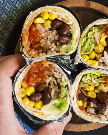 A hand is holding one of the four rainbow coloured round-shaped rolls from above. Inside red, yellow, brown, green and white pieces.