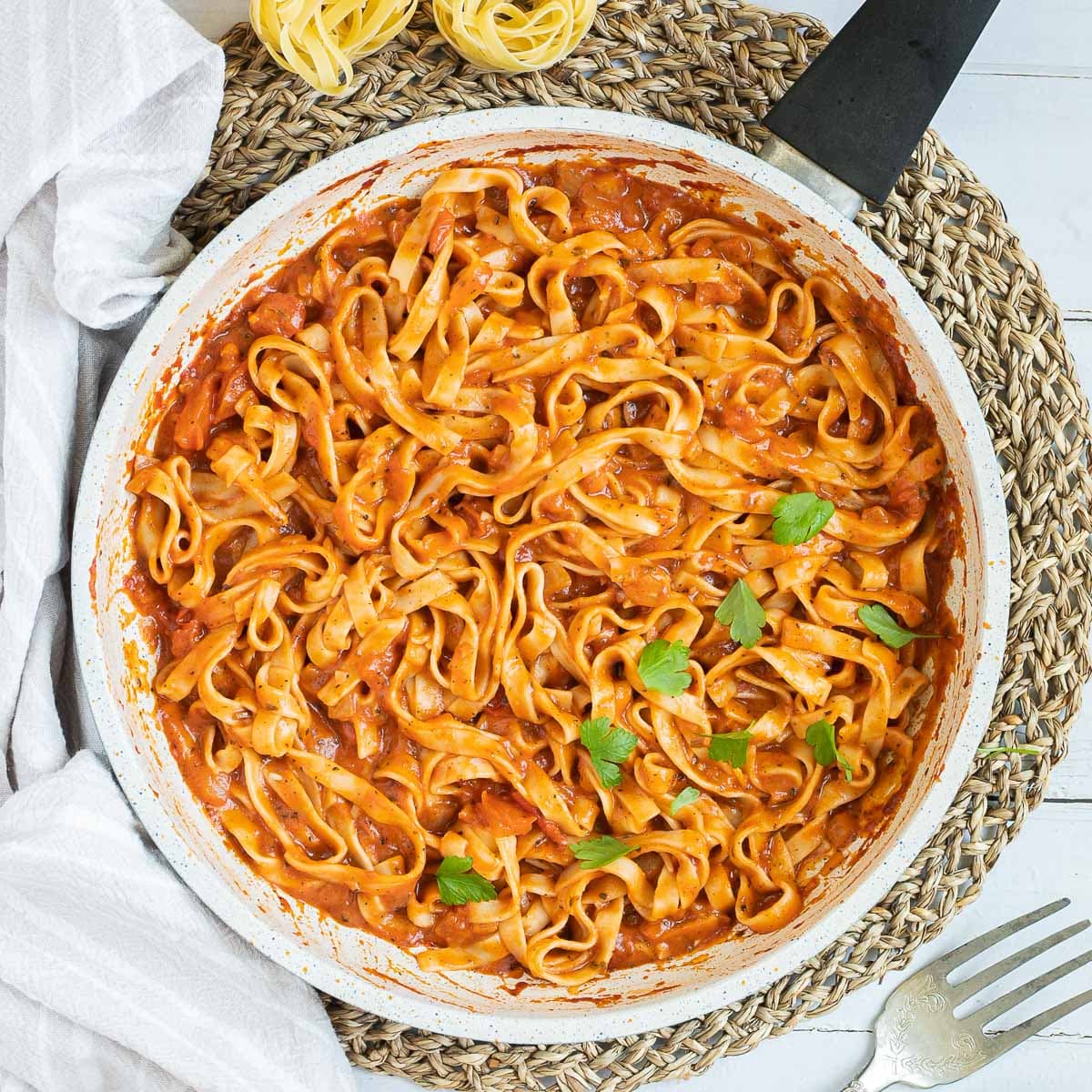Tagliatelle is mixed in and covered with a creamy light red tomato-based sauce topped with fresh green herbs in a white frying pan. Dry tagliatelle pasta and a cheese grater with parmesan cheese is next to it.