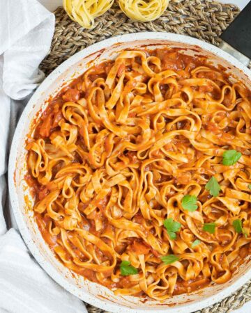 Tagliatelle is mixed in and covered with a creamy light red tomato-based sauce topped with fresh green herbs in a white frying pan. Dry tagliatelle pasta and a cheese grater with parmesan cheese is next to it.