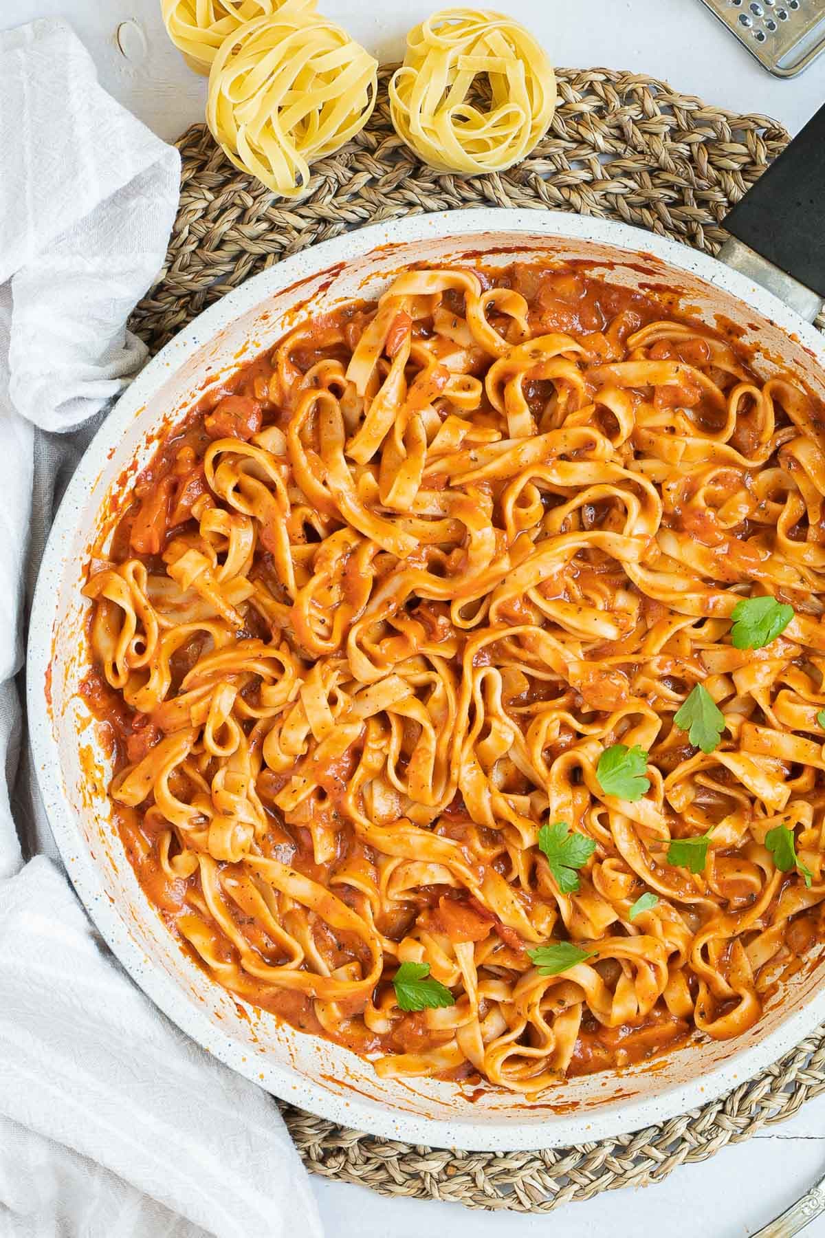 Tagliatelle is mixed in and covered with a creamy light red tomato-based sauce topped with fresh green herbs in a white frying pan. Dry tagliatelle pasta and a cheese grater with parmesan cheese is next to it.