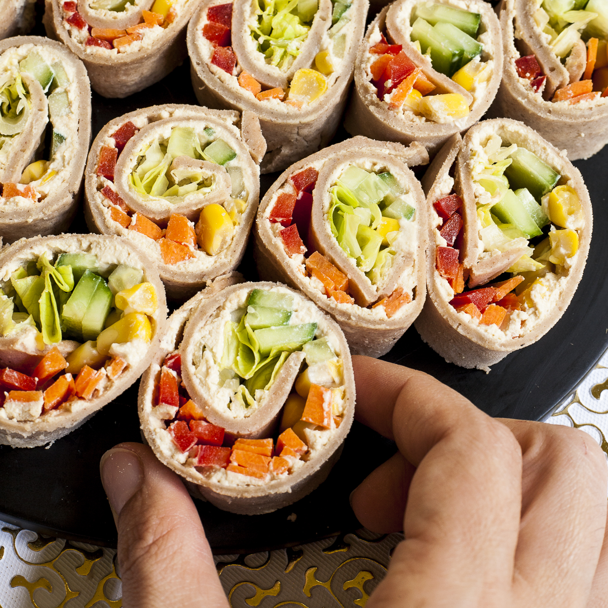 Colorful pinwheels of orange, red, yellow and green filling is placed closely on a black plate. A hand is grabbing one.