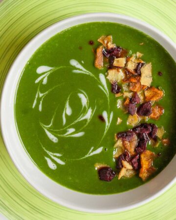 A bowl of green soup sprinkled with vegetable crisps and white swirls.