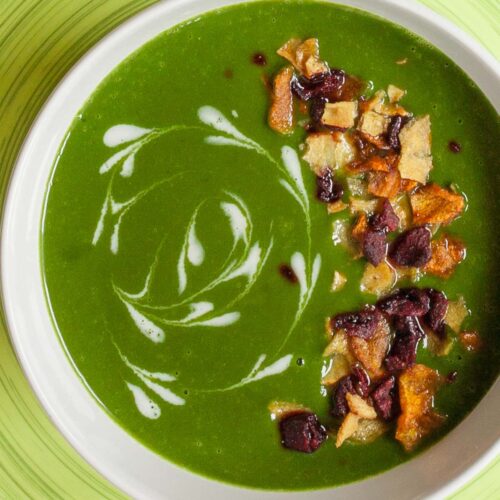 A bowl of green soup sprinkled with vegetable crisps and white swirls.