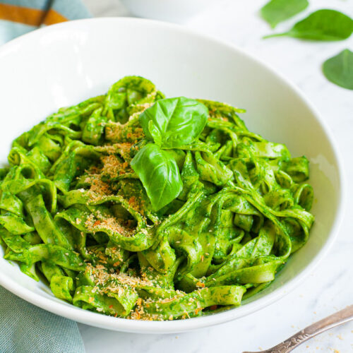White bowl with fettuccine pasta covered in green sauces sprinkled with yellow flakes and a pair of basil leaves. A fork is next to the bowl.