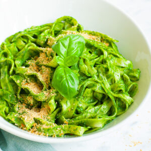 White bowl with fettuccine pasta covered in green sauces sprinkled with yellow flakes and a pair of basil leaves. A fork is next to the bowl.