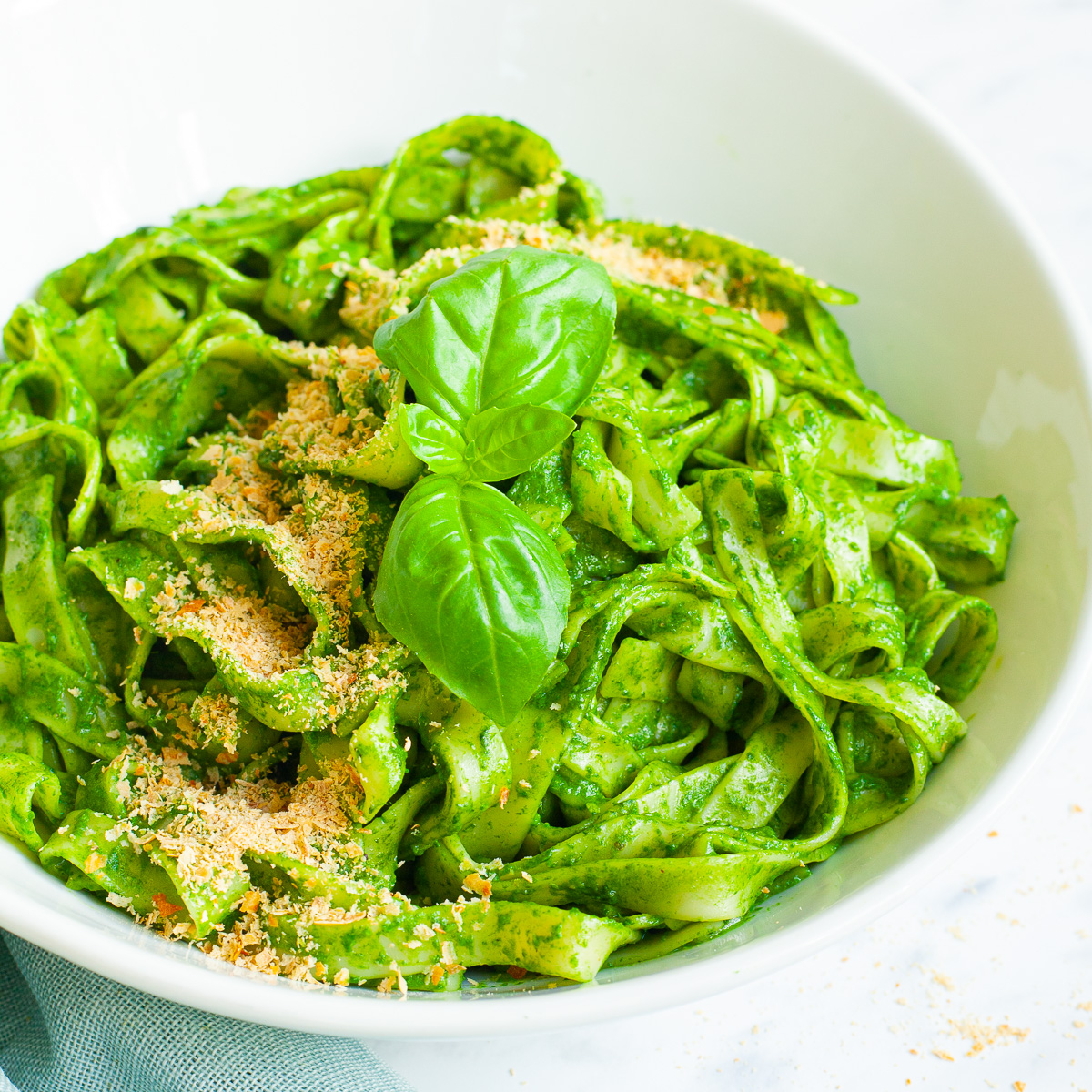 White bowl with fettuccine pasta covered in green sauces sprinkled with yellow flakes and a pair of basil leaves. A fork is next to the bowl.