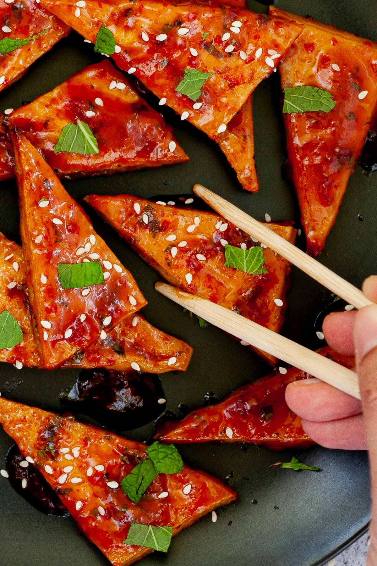 Triangle-shaped tofu slices in a red sticky glaze with freshly chopped green mint, sesame seeds on black plate. A hand with chopsticks is about to take one.