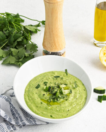 A white bowl with a vibrant green soup topped with freshly chopped herbs and cucumber pieces.