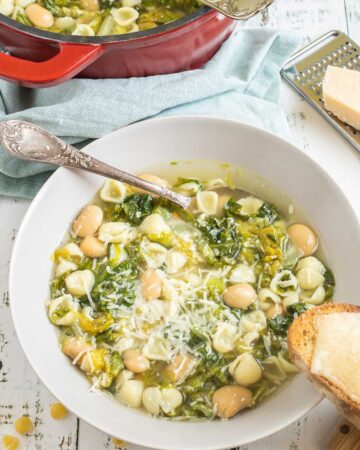 A white bowl of thin soup with white beans, chopped green escarole, small shell pasta, topped with grated cheese. Leftover soup is in a red enameled Dutch oven at the back. A cheesy, toasted bread is placed on the side of the bowl.